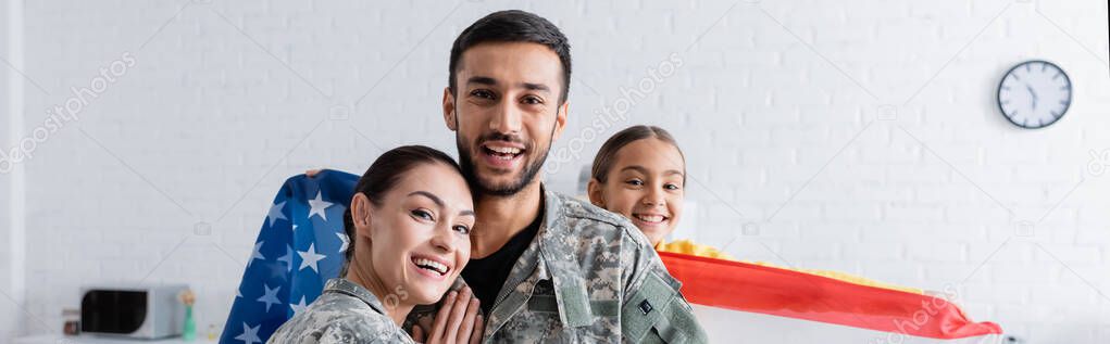 Smiling parents in military uniform looking at camera near child with american flag at home, banner 