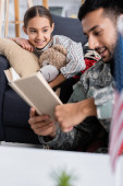 Smiling child with teddy bear looking at blurred father in military uniform reading book at home 