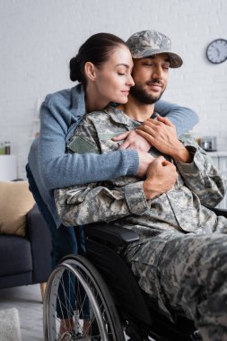 Woman hugging husband in camouflage uniform and wheelchair at home  clipart
