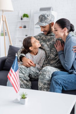 Memleketinde Amerikan bayrağı yanında eşine ve çocuğuna sarılan askeri üniformalı olumlu bir adam. 