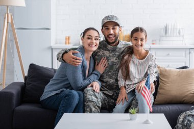 Kamuflaj üniformalı olumlu bir adam evdeki Amerikan bayrağının yanındaki koltukta ailesine sarılıyor. 
