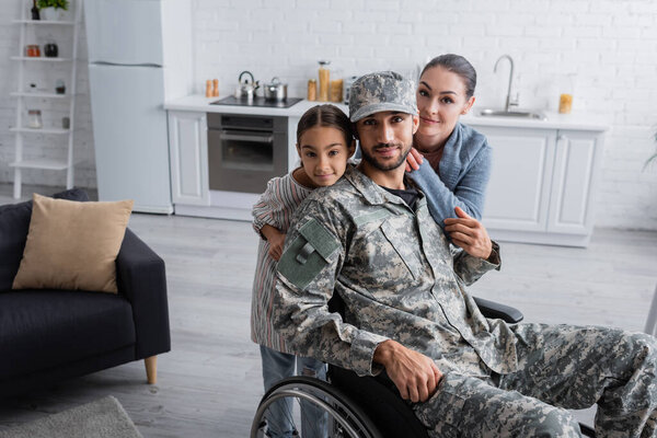 Мужчина в военной форме и инвалидной коляске смотрит в камеру рядом с семьей дома 