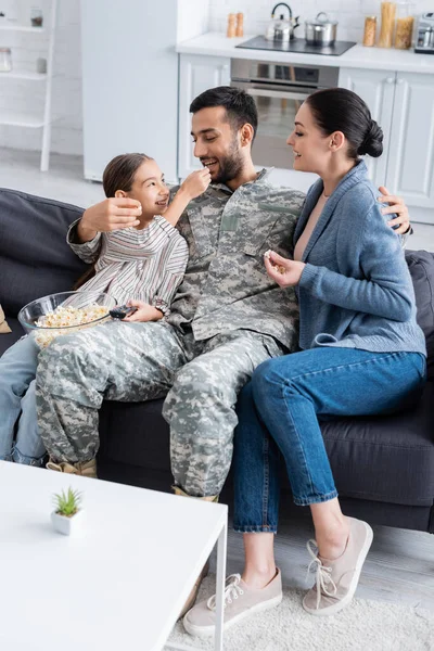小孩拿着爆米花和遥控装置靠近身穿军服的爸爸和坐在沙发上的妈妈 — 图库照片