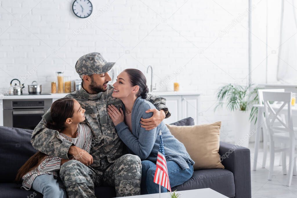 Man in camouflage uniform hugging family near american flag at home 