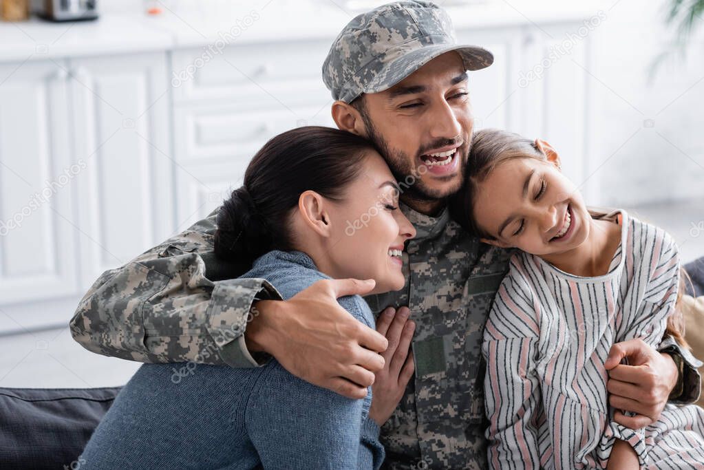 Joyful man in military uniform hugging daughter and wife at home 