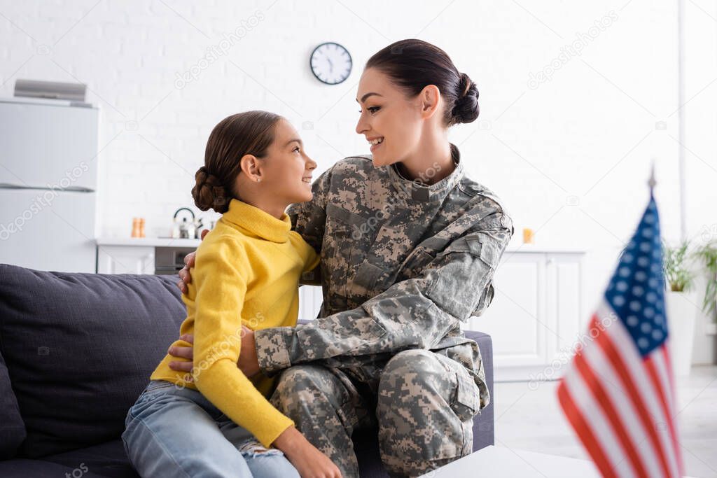 Side view of mom in military uniform hugging child near american flag on table at home 