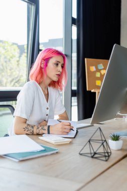 Not defterine pembe saçlı delinmiş bir iş kadını bilgisayar ekranına bakarken 