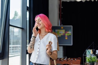 Pembe saçlı, gülümseyen iş kadını pencerenin yanında akıllı telefondan konuşuyor.