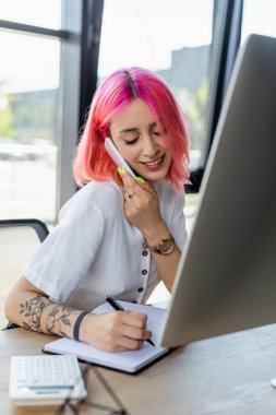 Pembe saçlı gülümseyen iş kadını akıllı telefondan konuşuyor ve monitörün yanındaki deftere yazı yazıyor. 