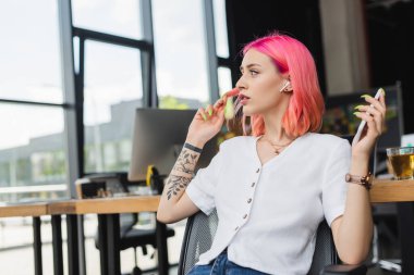 İşyerinde pembe saçlı ve kulaklıklı akıllı telefon tutan dövmeli bir iş kadını.