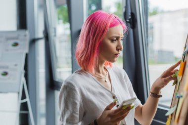 Pembe saçlı iş kadını akıllı telefon tutuyor ve tahtaya yapışkan notlarla işaret ediyor. 