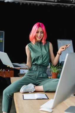 Pembe saçlı neşeli iş kadını dizüstü bilgisayarını tutuyor ve ofisteki monitöre bakıyor.