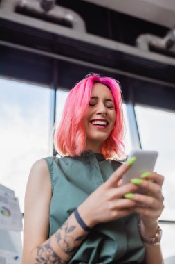 Akıllı telefon kullanan pembe saçlı, gülümseyen iş kadınının düşük açılı görüntüsü