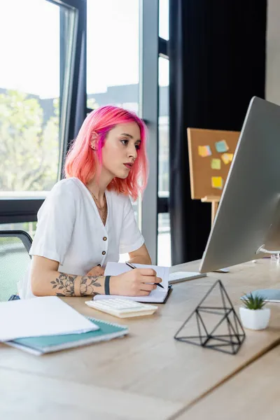 Przekłute Businesswoman Różowymi Włosami Pisanie Notebooku Podczas Patrząc Monitor Komputera — Zdjęcie stockowe
