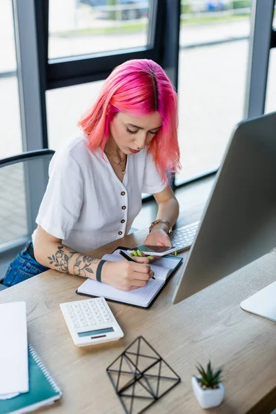 Młoda Bizneswoman Różowymi Włosami Pisanie Notebooku Trzymając Smartfona — Zdjęcie stockowe