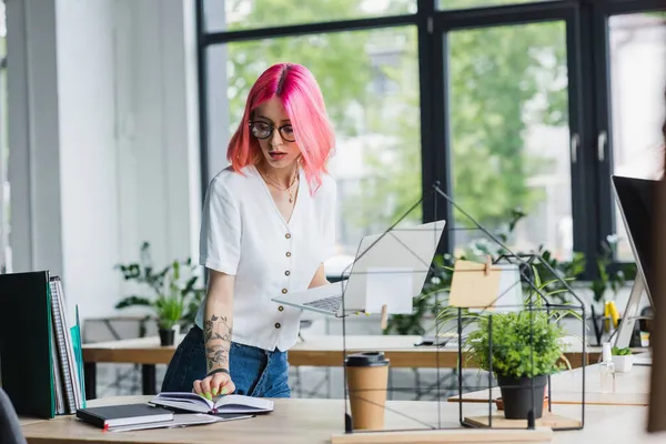 Młoda Businesswoman Różowymi Włosami Trzyma Laptopa Patrząc Notatnik — Zdjęcie stockowe