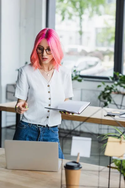 Genomborrad Affärskvinna Med Rosa Hår Håller Anteckningsbok Och Pekar Bärbar — Stockfoto