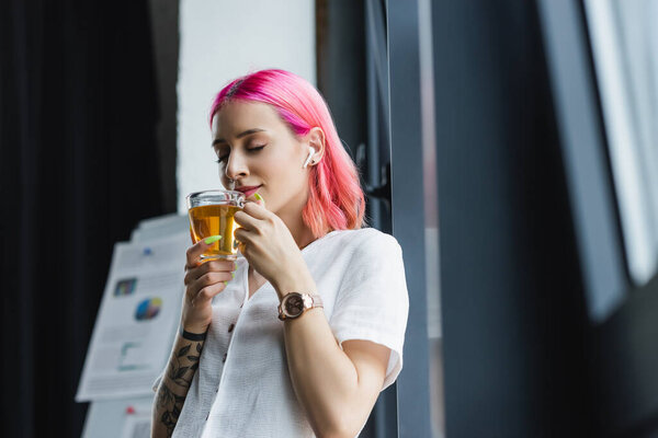 pleased businesswoman with pink hair and earphone smelling cup of tea in office