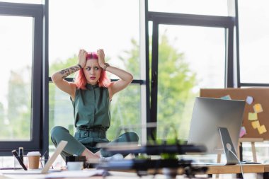 stressed businesswoman with pink hair sitting with crossed legs on desk in office clipart
