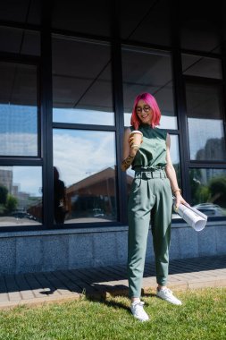full length of pleased businesswoman with pink hair holding rolled paper and coffee to go outside  clipart