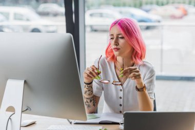 Ofiste bilgisayar monitörünün yanında pembe saçlı gözlüklü bir iş kadını.