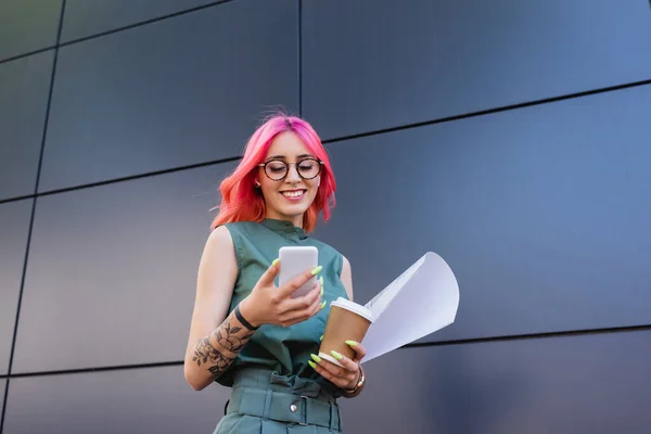 happy and tattooed businesswoman in earphones using smartphone while holding paper cup