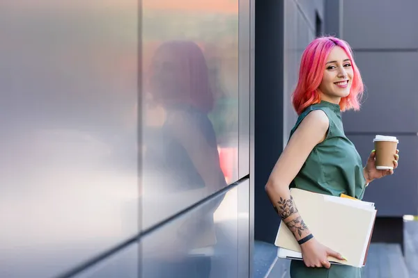 tattooed and happy businesswoman with pink hair holding folder, laptop and coffee to go outside