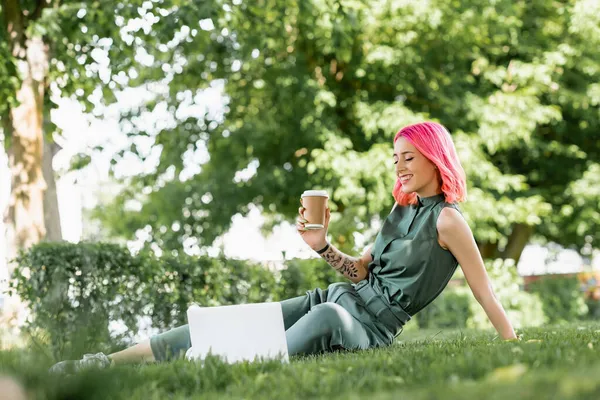 芝の上にノートパソコンの近くにピンクの髪の保持紙カップを持つ陽気な若い女性 — ストック写真