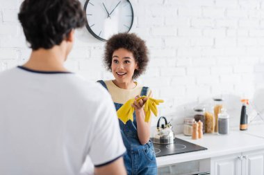 Plastik eldivenli gülümseyen Afro-Amerikan kadın mutfakta bulanık bir adama bakıyor. 