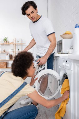 Kıvırcık Afrikalı Amerikalı kadın çamaşır makinesini erkek arkadaşının yanına kirli çamaşır sepetiyle dolduruyor. 