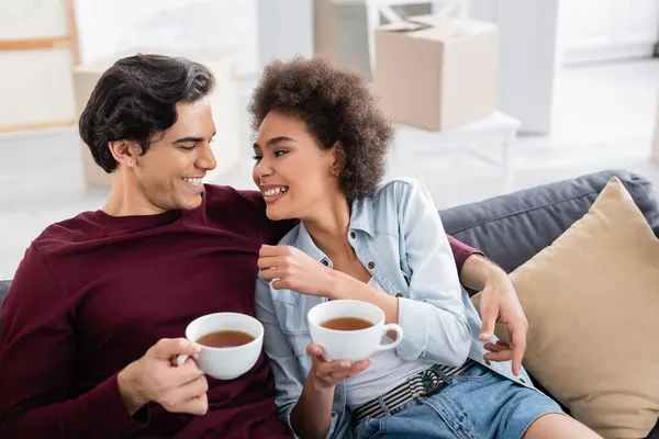 Alegre Pareja Multiétnica Sosteniendo Tazas Mientras Descansa Sofá — Foto de Stock