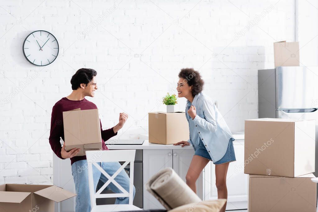 excited interracial couple celebrating relocation near carton boxes