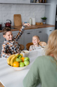 usmívající se chlapec ukazuje prstem na bratra při snídani s cereáliemi a čerstvé ovoce ner rozmazané maminka