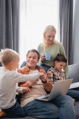 Kocasının yanında çay içen mutlu bir kadın laptopuyla kanepesinde çocuklarla eğleniyor.