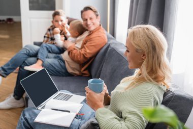 Not defteri, dizüstü bilgisayarı ve çay bardağı olan bir kadın. Bulanık bir ailenin yanında, koltukta oturuyor.