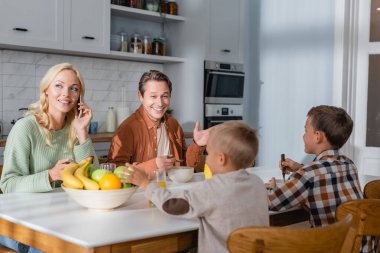 Karısı cep telefonundan ararken neşeli bir adam kahvaltıda oğullarıyla konuşuyor.