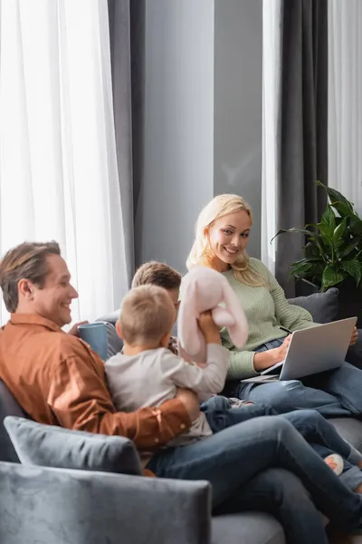 Sorrindo Mulher Trabalhando Laptop Sofá Enquanto Marido Brincando Com Filhos — Fotografia de Stock