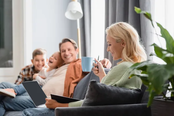 一个微笑的女人 带着一杯茶 带着笔记本电脑和笔记本在沙发上与模糊的家人一起工作 — 图库照片