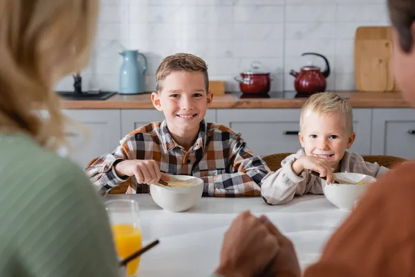 Usmívající Kluci Jíst Lahodné Kukuřičné Vločky Blízkosti Rozmazané Rodiče Kuchyni — Stock fotografie