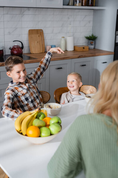 smiling boy pointing with finger at brother during breakfast with cereals and fresh fruits ner blurred mom