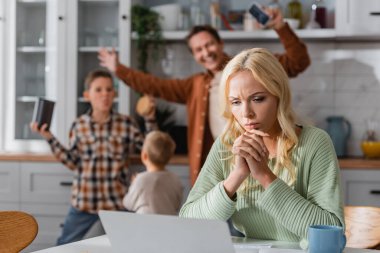 Bulanık aile, mutfakta dizüstü bilgisayarın yanında çalışan düşünceli kadının yanında eğleniyor.