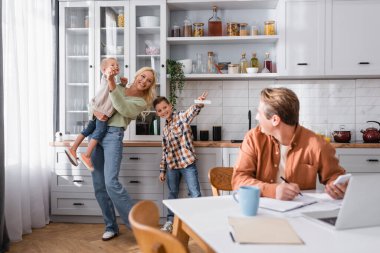 cheerful woman having fun with sons near blurred husband working in kitchen clipart