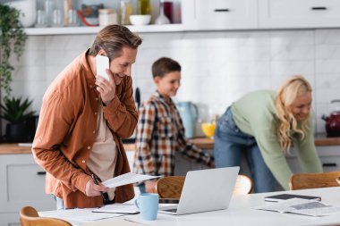 Mutfaktaki bulanık ailenin yanında doküman ve laptopla çalışırken akıllı telefondan konuşan mutlu bir adam.