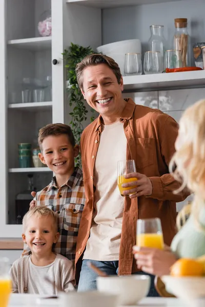 息子の近くで笑うオレンジジュースとキッチンでぼやけた妻を持つ陽気な男 — ストック写真