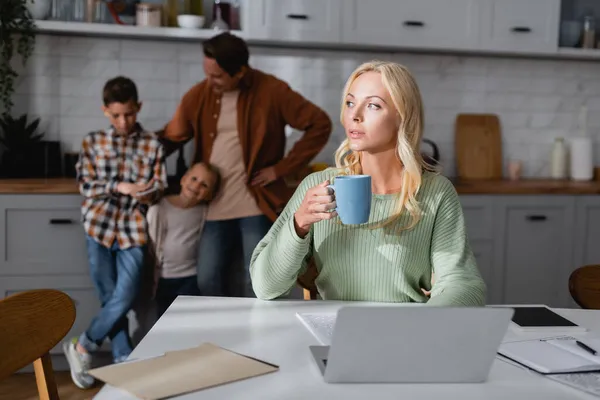 Thoughtful Woman Cup Tea Sitting Laptop Documents Blurred Husband Kids — Stock Photo, Image