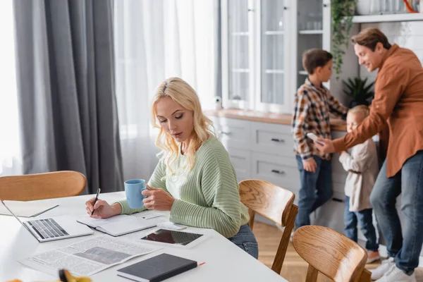 Woman Cup Tea Writing Notebook Gadgets Blurred Husband Taking Selfie — Stock Photo, Image