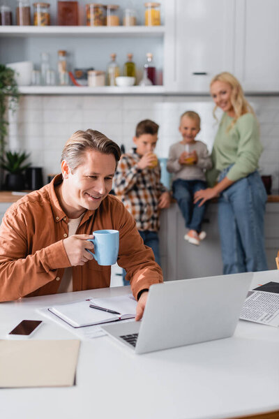 smiling man with cup of tea working in kitchen near laptop and  family on blurred background