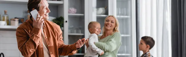 キッチンやバナーの中のぼやけた妻と息子の近くのスマートフォンで話している笑顔の男 — ストック写真