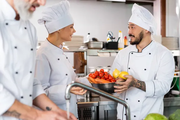 Sonriente Asiático Chef Celebración Bowl Con Verduras Cerca Colegas Grifo — Foto de Stock