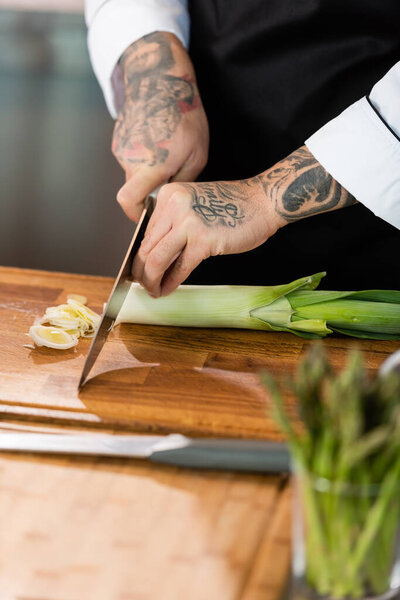 Cropped view of tattooed chef cutting leek near asparagus on blurred foreground in kitchen 